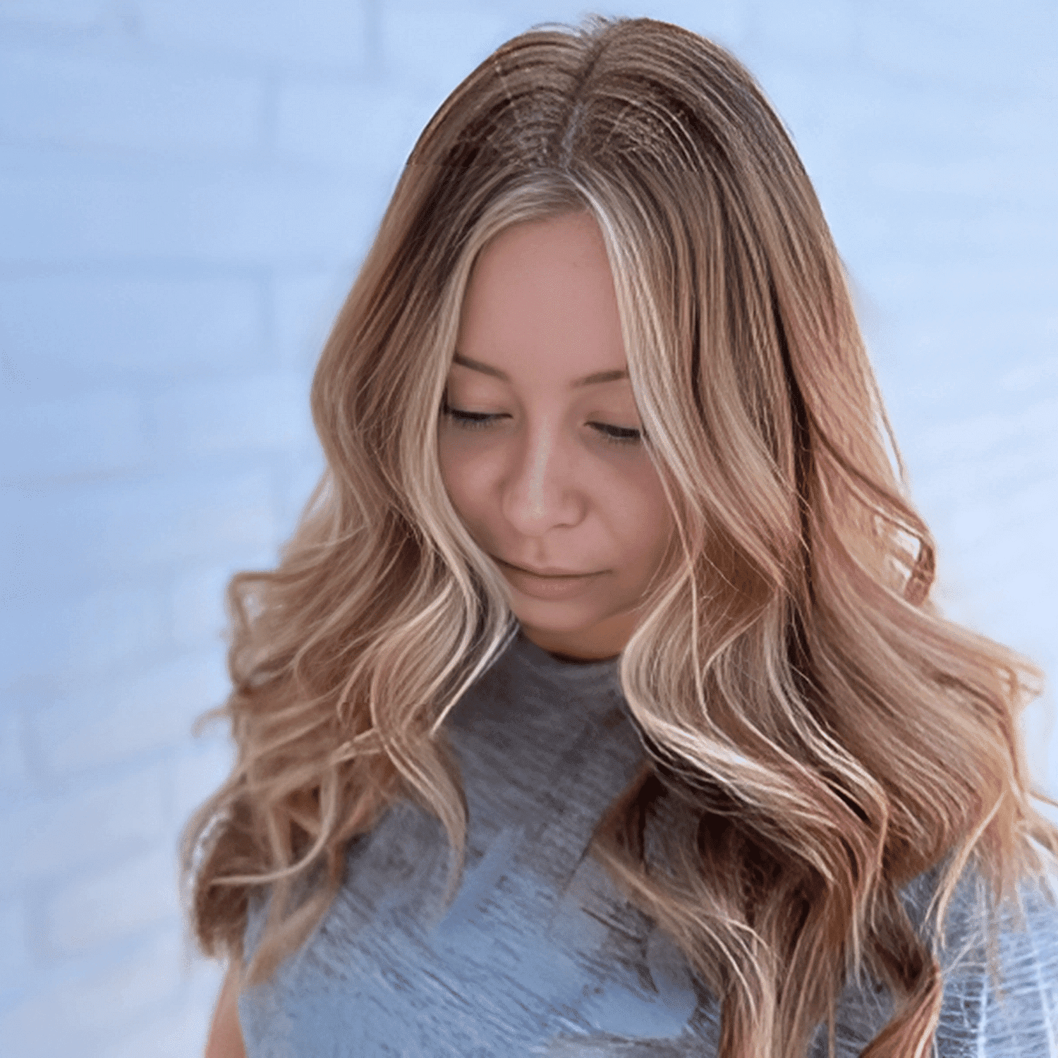 Woman with wavy, highlighted hair against a blurred brick wall background.